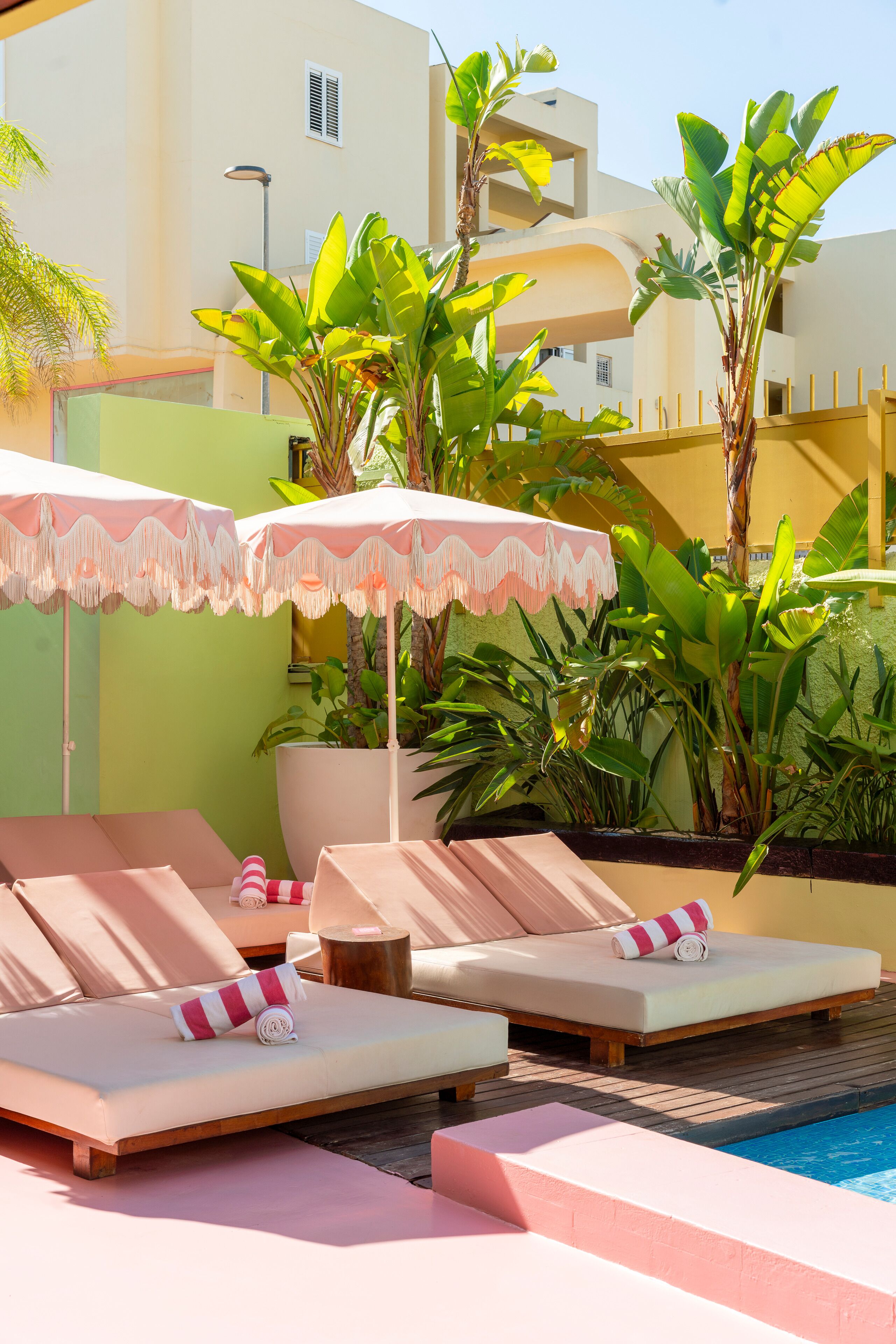 A poolside area with lounge chairs and umbrellas, surrounded by lush greenery and a modern building in the background.