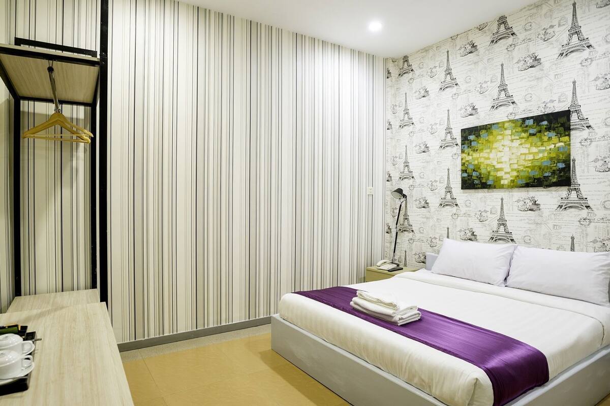 Ein Hotelzimmer mit Bett, Schreibtisch mit Stuhl, einer Garderobe, einem Nachttisch mit Lampe und einer Wand mit Eiffelturm-Tapete.