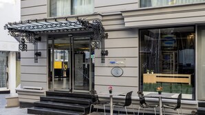 Een hotelentree met een glazen deur, een buitenzitgedeelte met twee tafels en vier stoelen, en een klein tuinzicht door het raam.