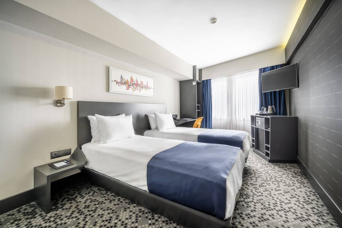 A hotel room with two beds, a desk, a TV, and a large window with curtains.
