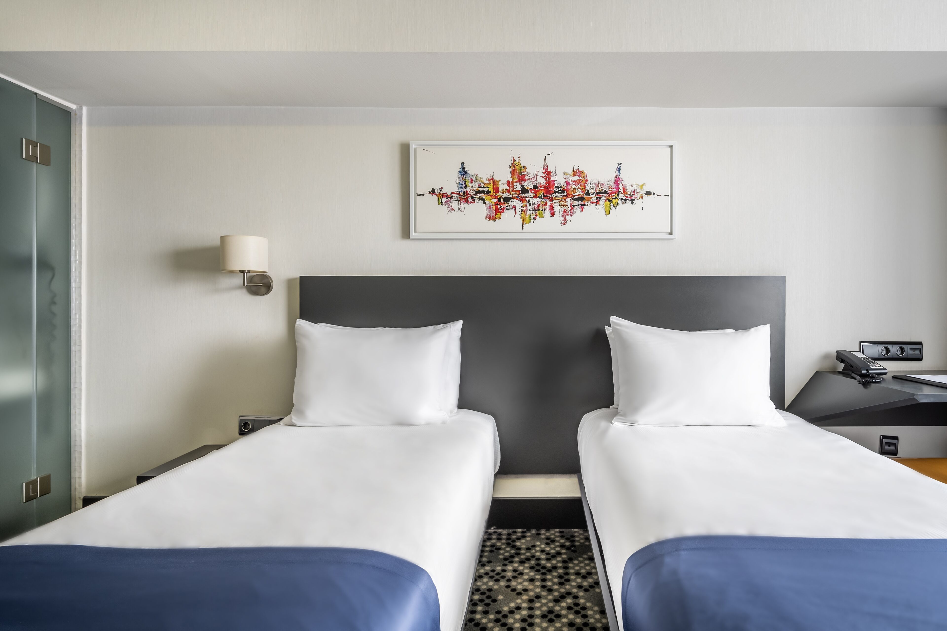 Hotelkamer met twee enkele bedden, een grijze hoofdbord, een aan de muur bevestigd kunstwerk, een nachtkastje met lamp en een klein bureau met telefoon en radio.