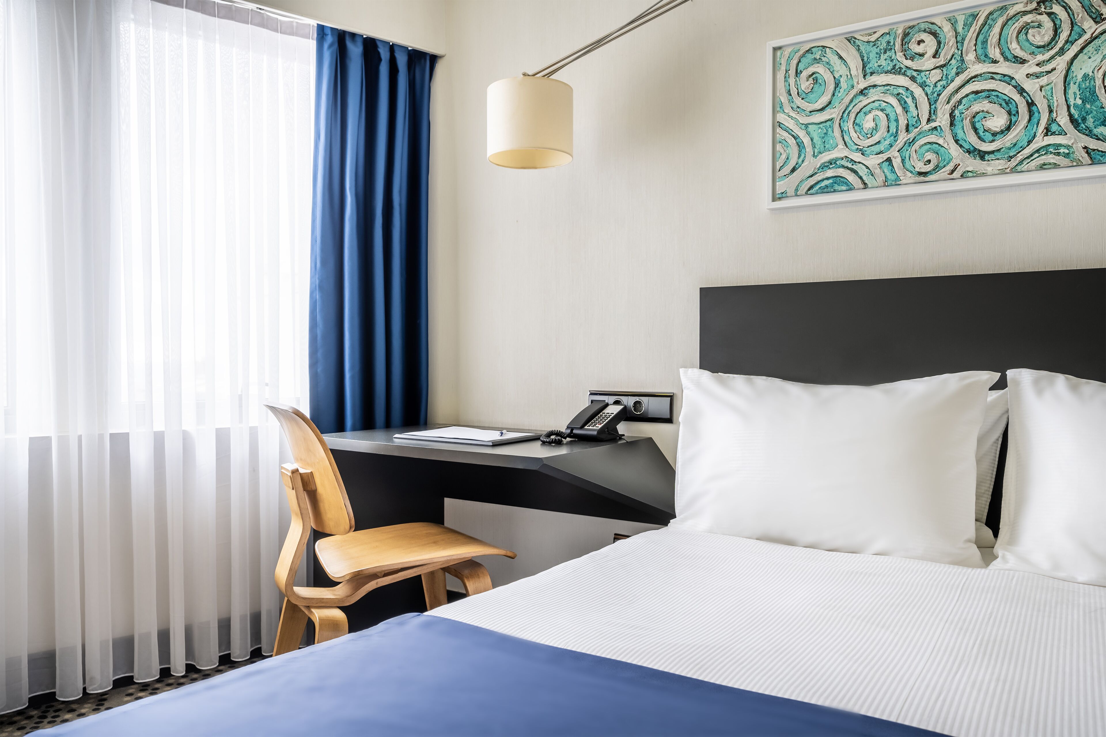 Ein Hotelzimmer mit einem Bett, einem Schreibtisch mit Telefon, einem Sessel und einem Fenster mit Vorhängen.