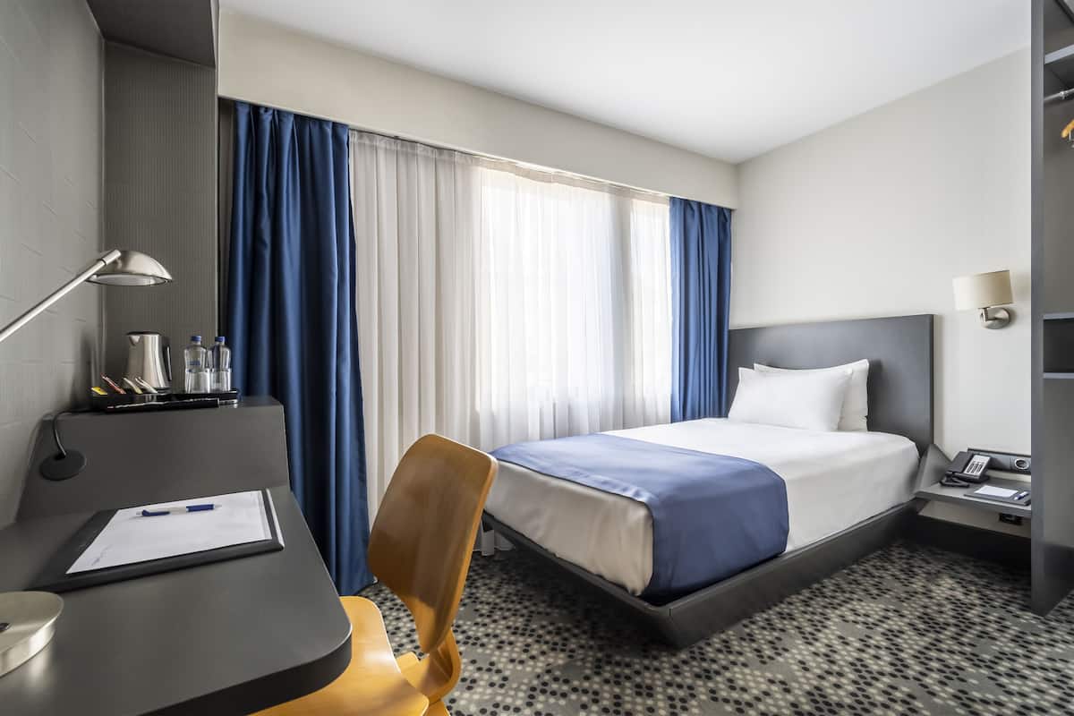 A hotel room with a bed, desk, chair, lamp, and a nightstand with a phone.