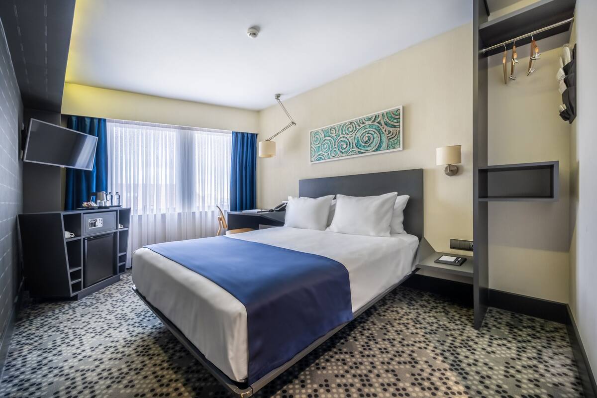 A hotel room with a bed, a desk, a TV, a painting, and a coat rack.