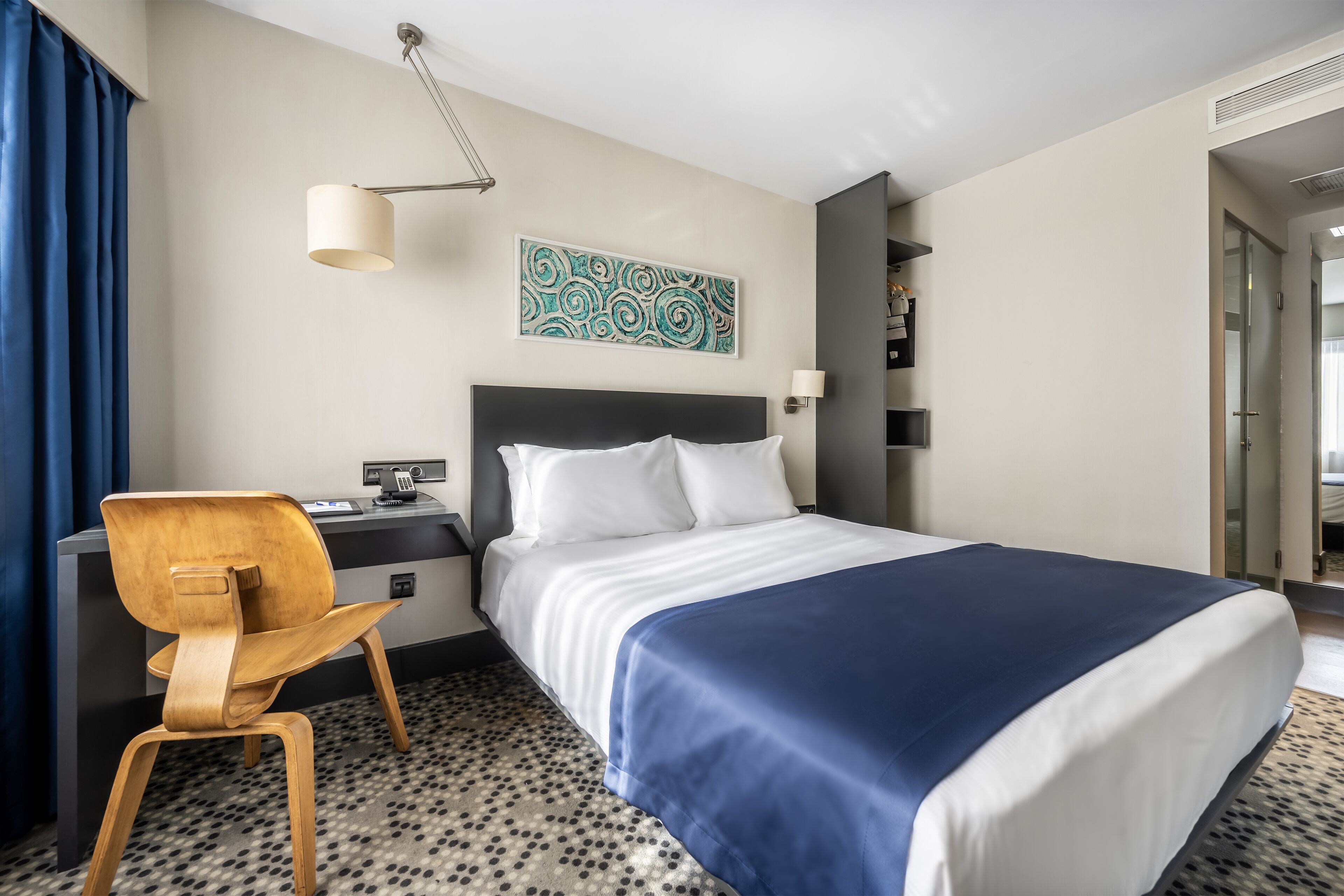 A hotel room with a bed, a desk, a chair, a wall art piece, and a patterned carpet.