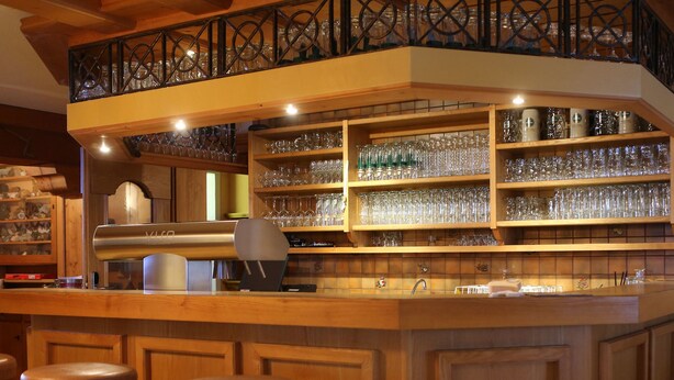 Una zona de bar con estantes de madera llenos de vasos y botellas, una barra y dos taburetes.
