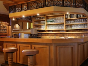 Una zona de bar con estantes de madera llenos de vasos y botellas, una barra y dos taburetes.