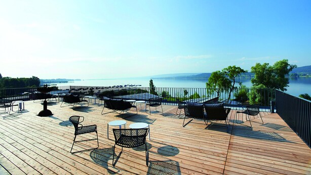 A wooden deck with outdoor seating, a view of the water, and a distant shoreline. - Hotel Bora HotSpaResort (Radolfzell am Bodensee)