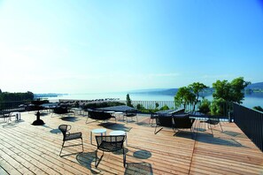A wooden deck with outdoor seating, a view of the water, and a distant shoreline. - Hotel Bora HotSpaResort (Radolfzell am Bodensee)