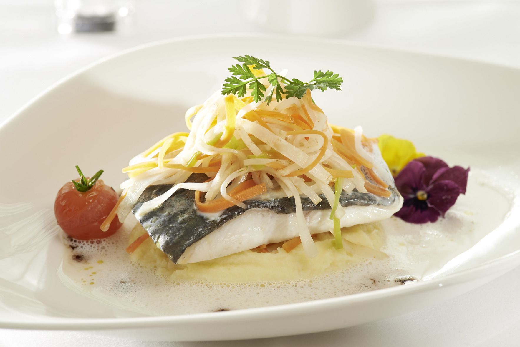En vit tallrik med en fiskfilé, strimlad grönsallad, en körsbärstomat och en lila blomma.