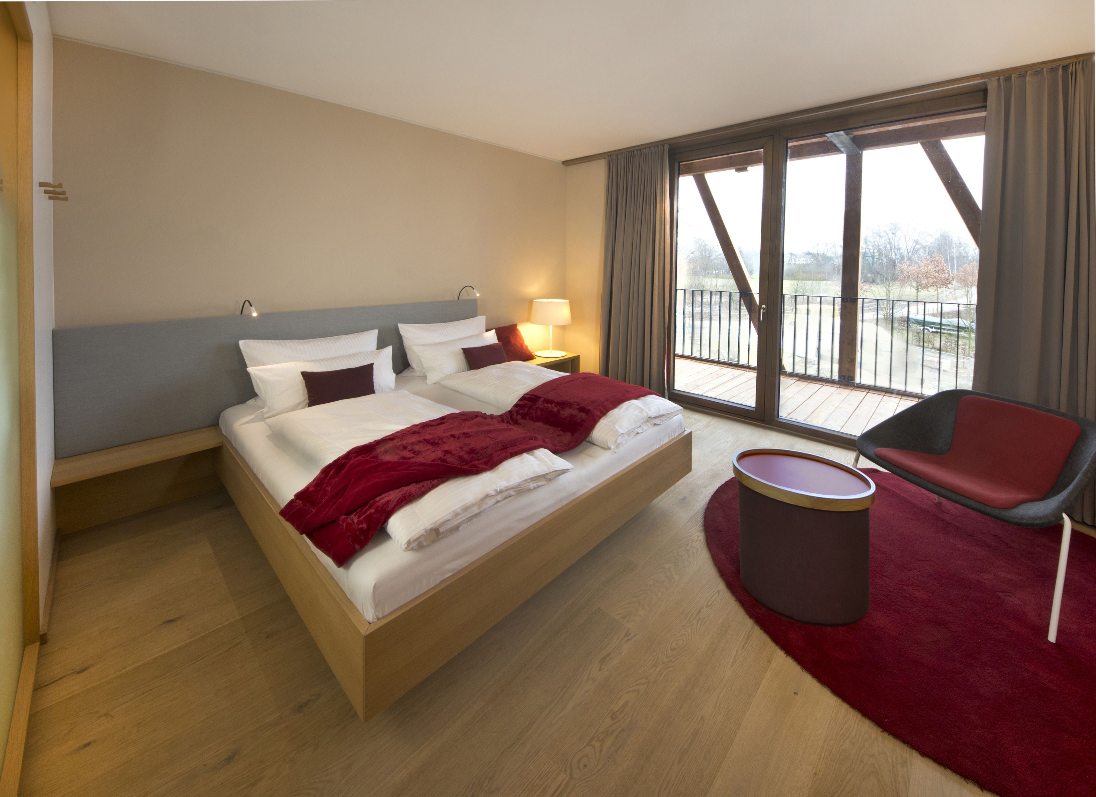A hotel room with a large bed, a chair, a small table, and a view of the outdoors through a large window.