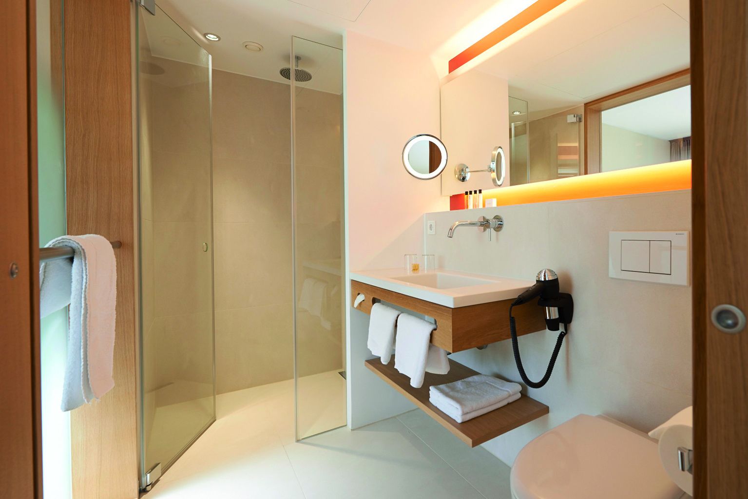 Ein modernes Badezimmer mit einer in Glas eingefassten Dusche, einem hölzernen Waschtisch mit Waschbecken und einem Spiegel mit einem Ringlicht.
