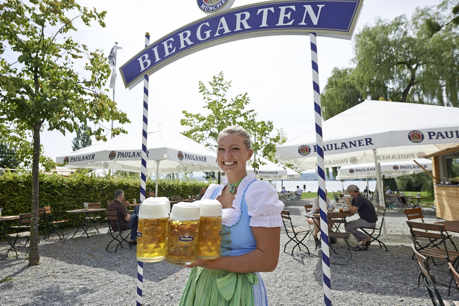 Eine Frau hält drei Bierkrüge vor einem Schild, auf dem 'Biergarten' steht.