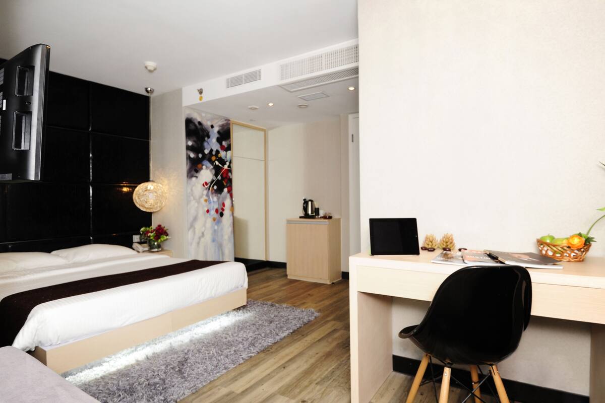 Un dormitorio moderno con una cama grande, un escritorio y una silla. Hay un cuadro en la pared, un mueble pequeño y una cesta de fruta en el escritorio.