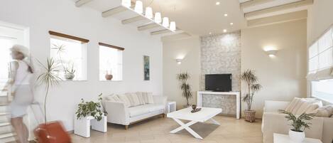 Un salón espacioso con un sofá blanco, una mesa de centro y un televisor. Hay plantas en macetas y una chimenea.