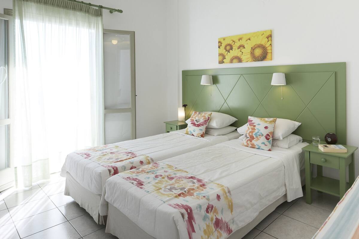 Un llit doble amb roba de llit floral, un panell capçal verd, dues taules de nit amb làmpades i una obra d'art amb girasols a la paret.