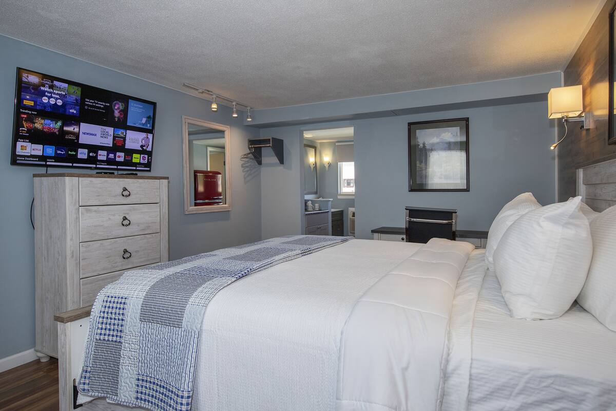 Ein Schlafzimmer mit einem großen Bett, einem an der Wand befestigten Fernseher, der verschiedene Sender zeigt, einer Kommode und einem Spiegel.