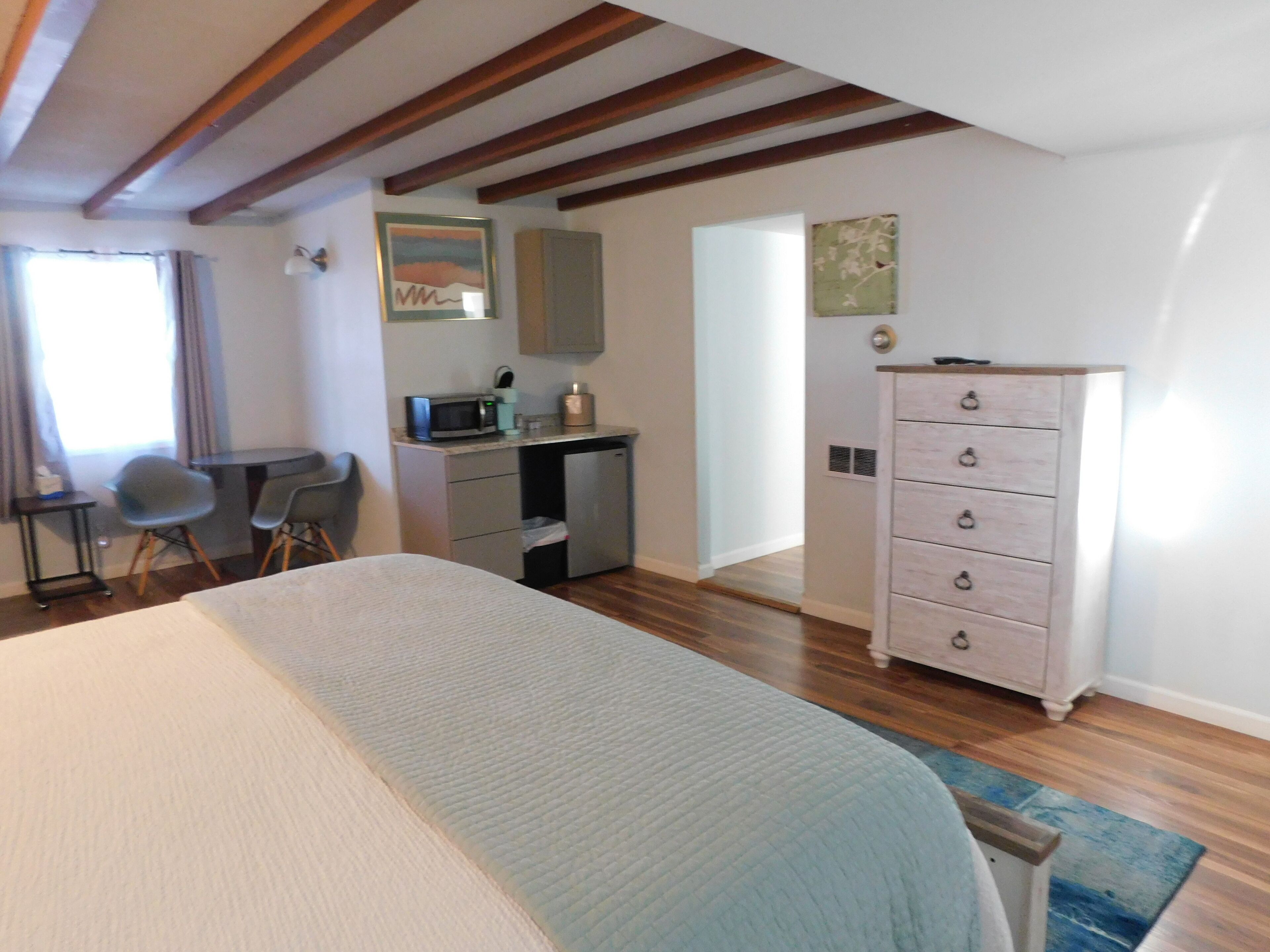 A bedroom with a bed, a desk, a chair, a dresser, and a kitchenette.