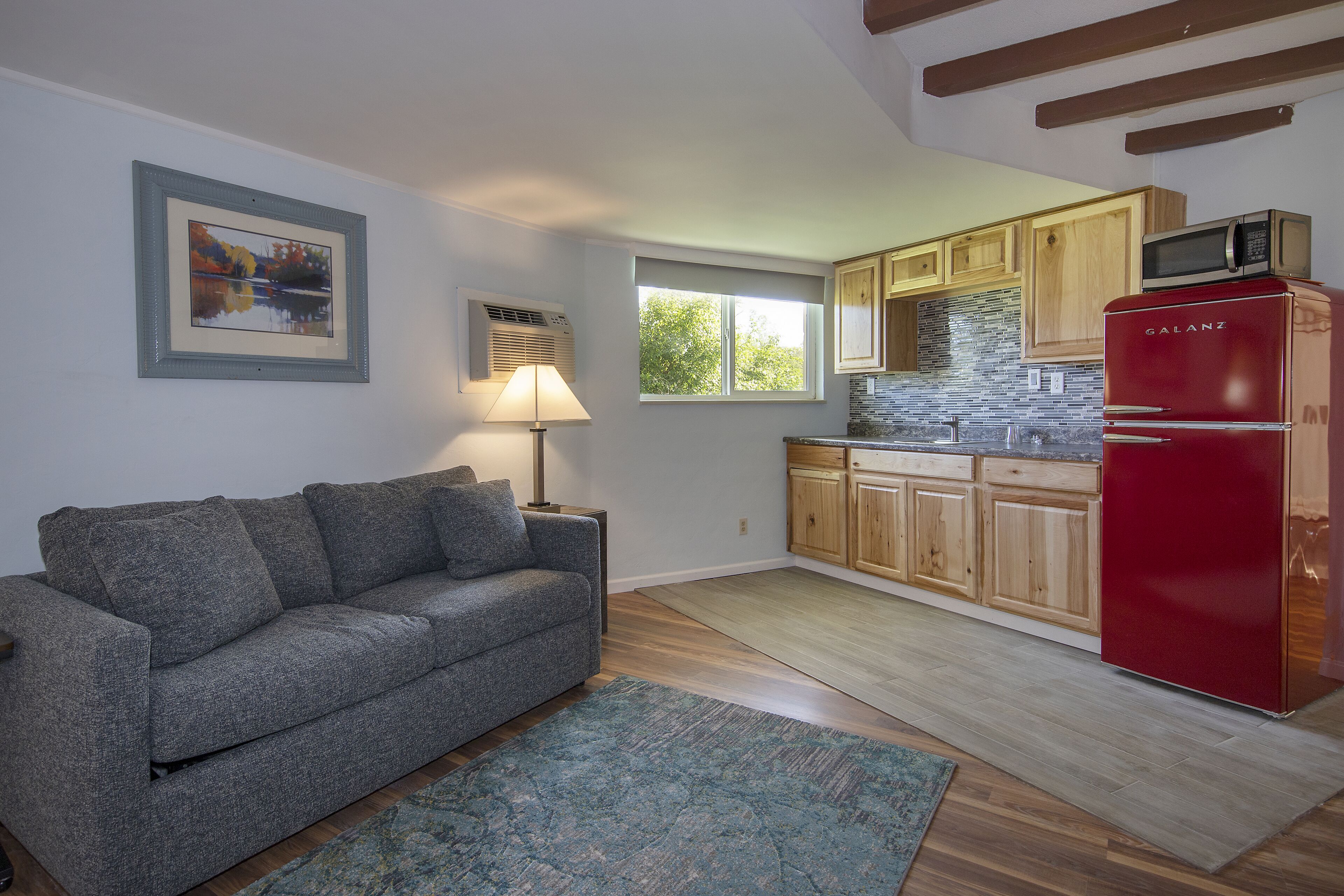 Un espacio habitable compacto con sofá gris, refrigerador rojo, armarios de madera, una ventana y un cuadro enmarcado en la pared.