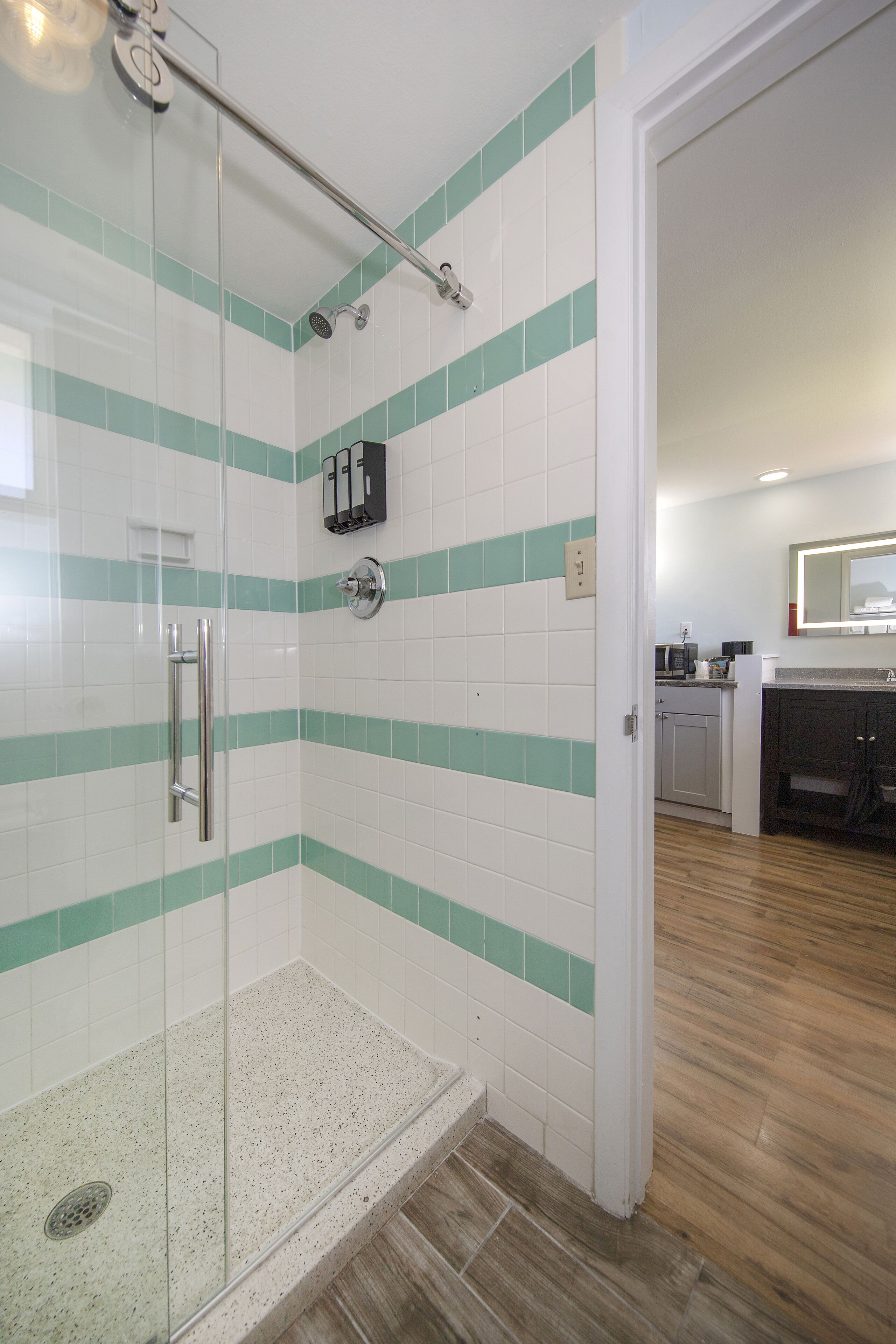 Un área de ducha con puerta de vidrio, pasamanos de metal y pared revestida con azulejos blancos y verdes.