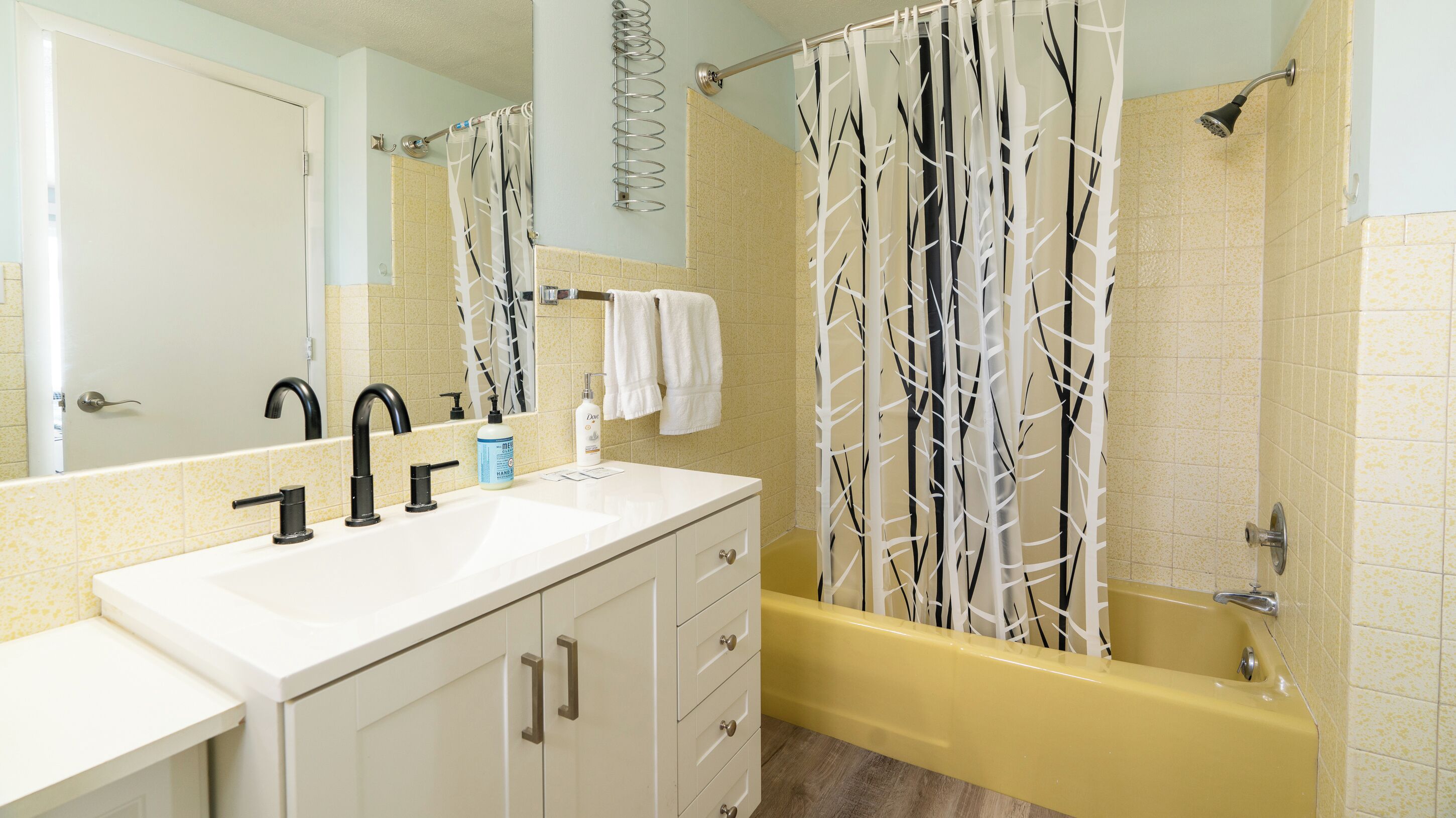 Un baño con un lavamanos blanco, grifos negros, una bañera amarilla y una cortina de ducha con estampado de árboles.