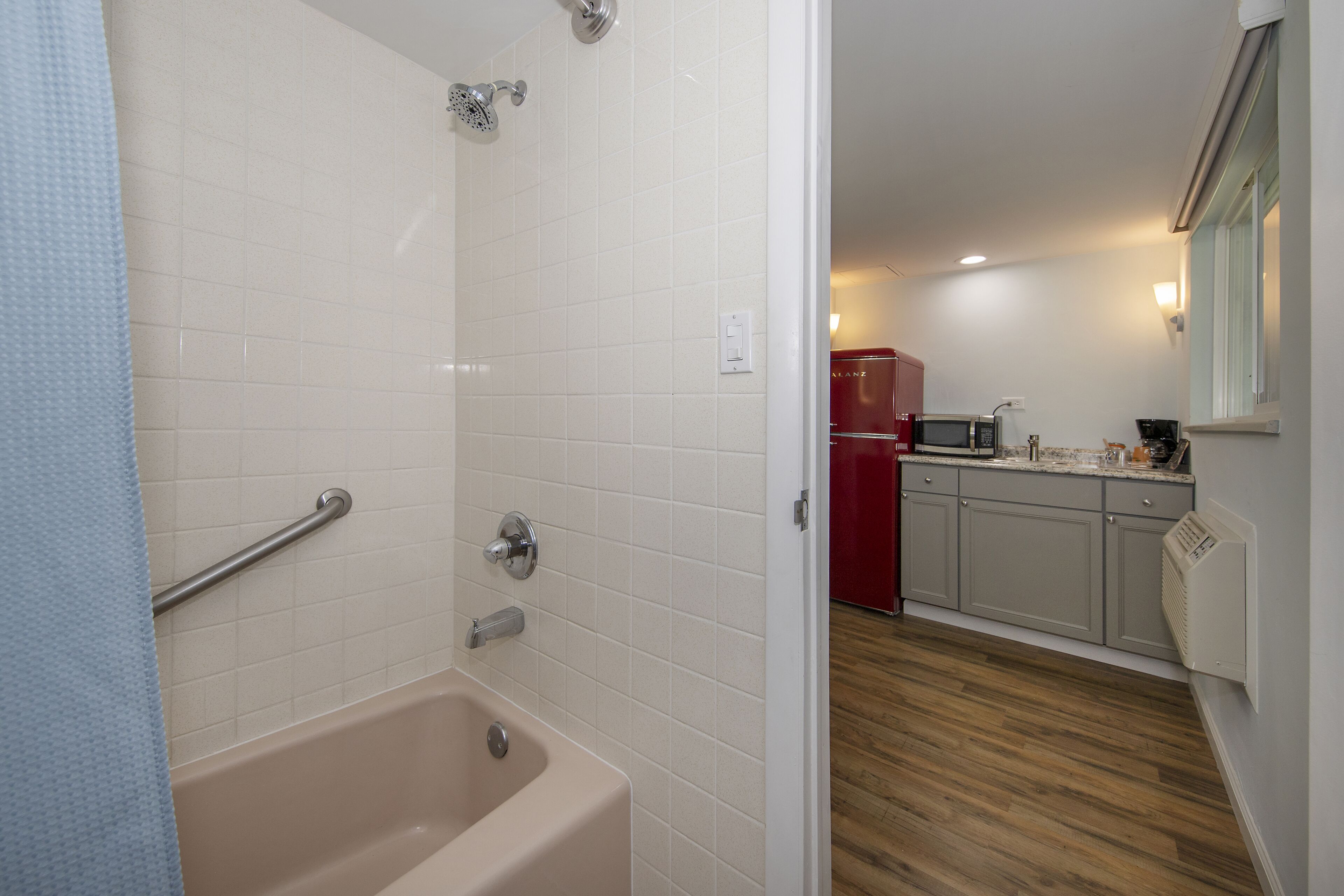 A bathroom with a bathtub, a shower, and a toilet. There is a wooden floor and a kitchen area visible in the background.