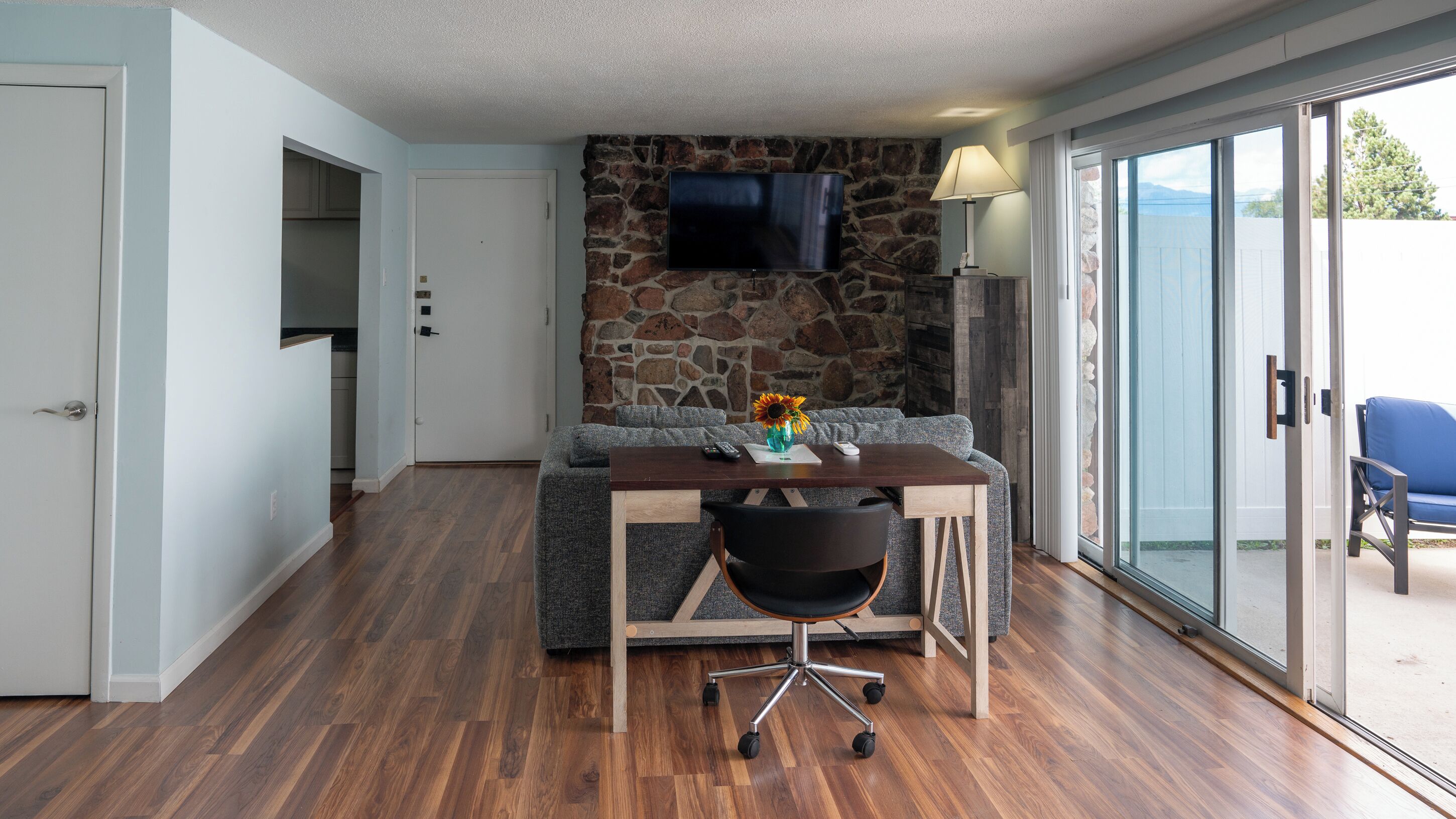 Een kamer met een stenen muur, een flatscreen televisie boven een houten tafel, een bank en een stoel met wielen.