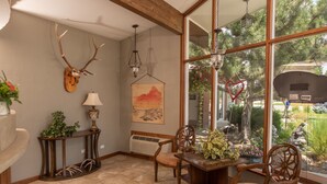 A dining area with a wooden table and chairs, a wall-mounted lamp, a framed painting, and a large window with a view of greenery and a parked vehicle. - Silver Ridge Lodge (Salida)