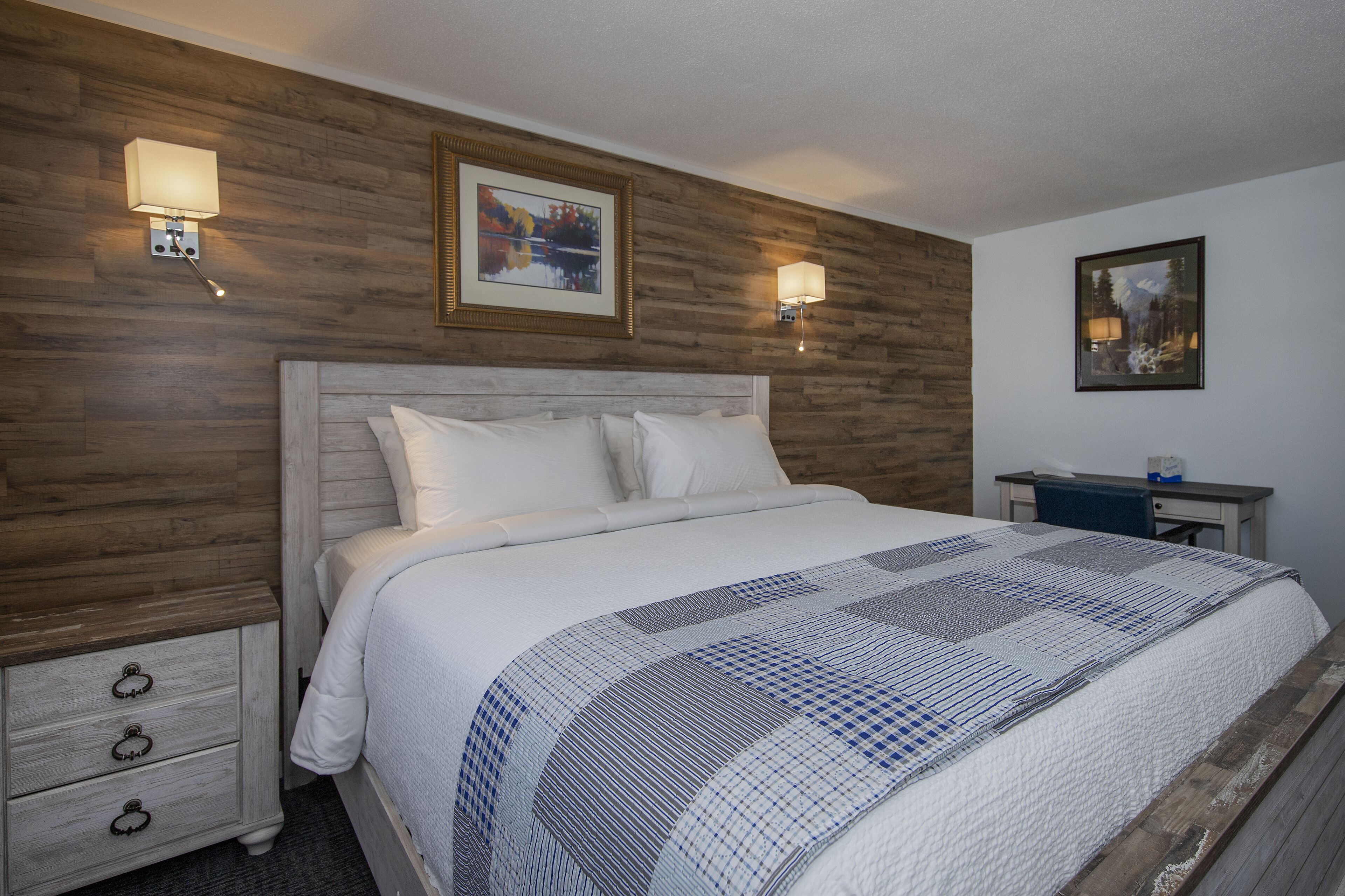 A neatly made bed with a quilt, wooden panel walls, two wall-mounted lamps, and a framed picture above the bed.