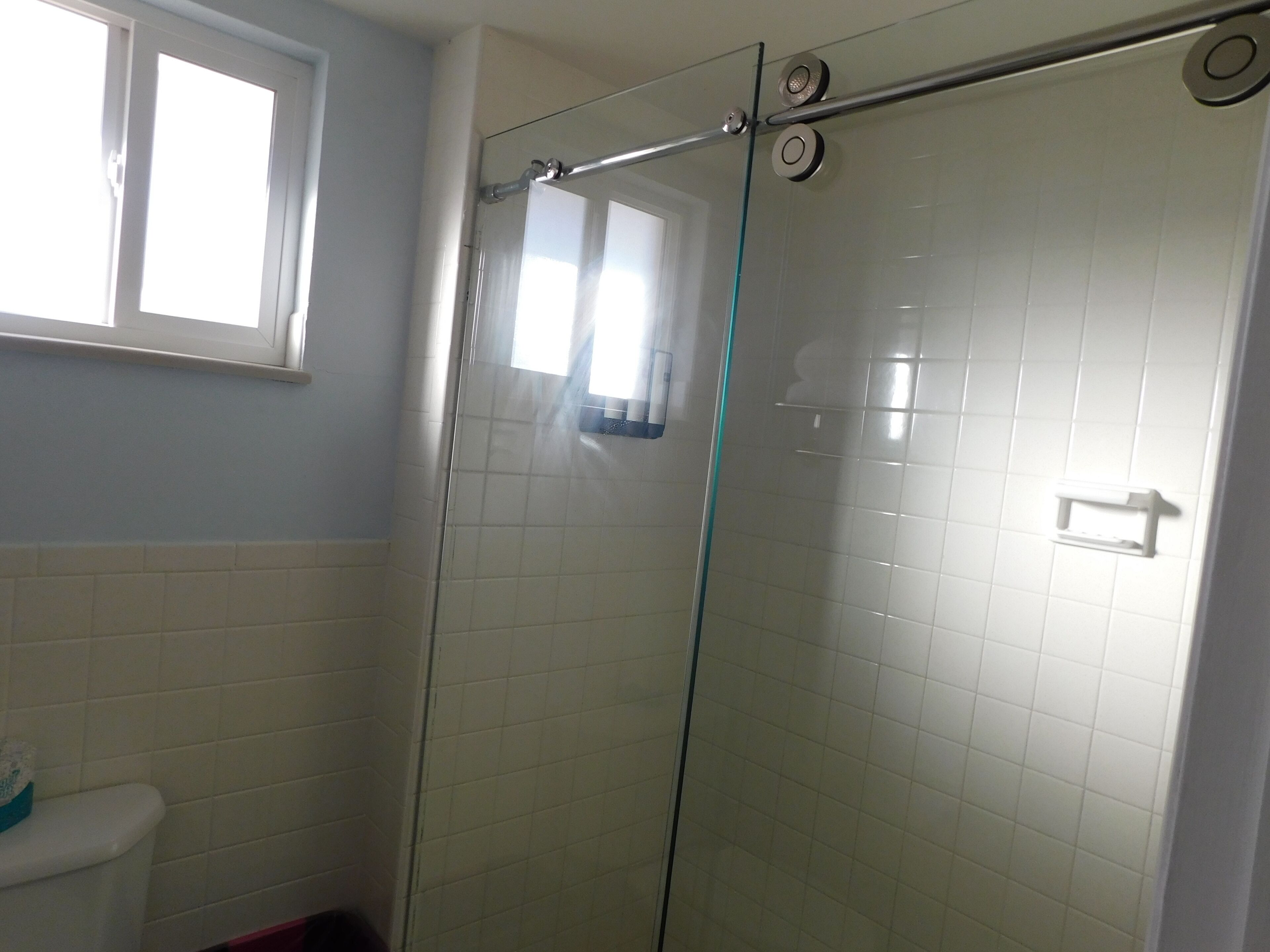 A bathroom with a glass shower enclosure, white tiles, and a window above the toilet.