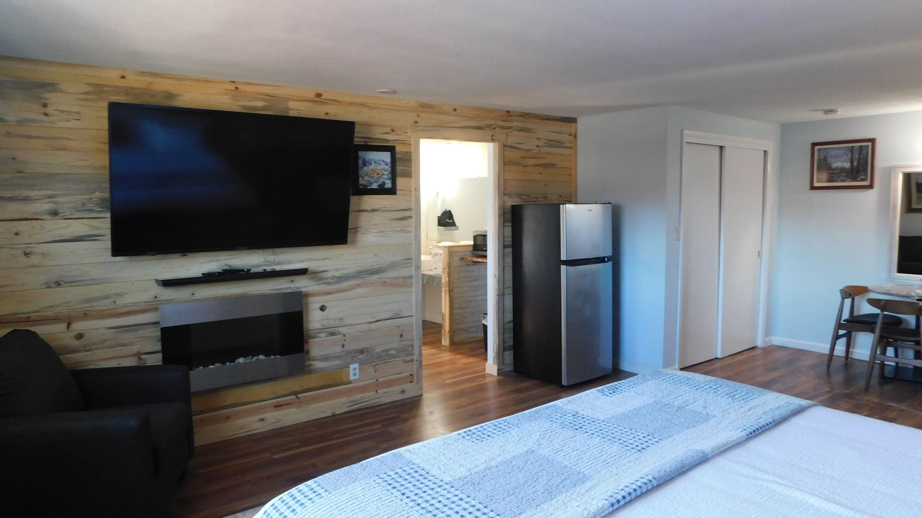 Een moderne kamer met een flatscreen televisie aan een houten wand, een koelkast, een eethoek met een tafel en stoelen, en een groot bed.