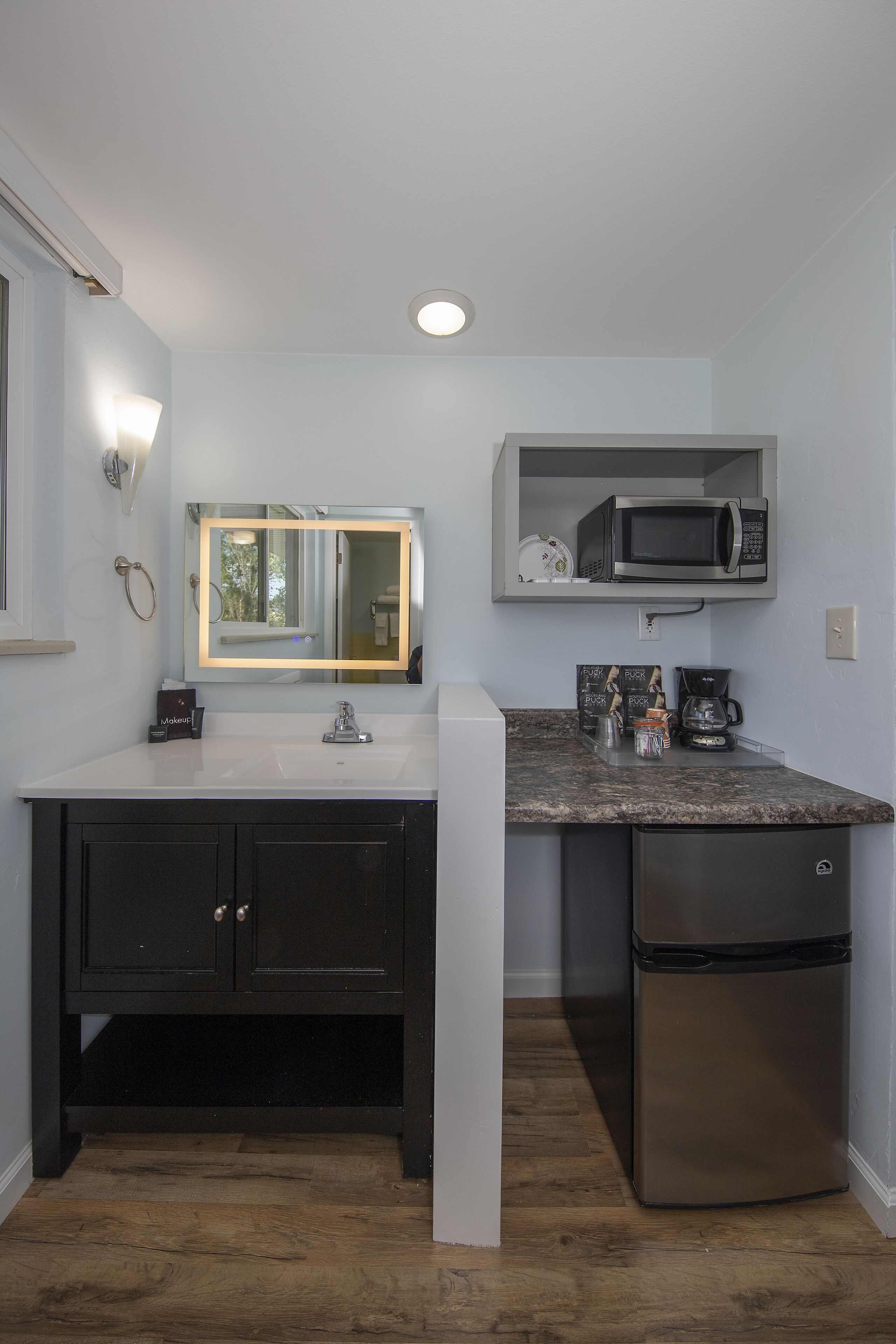 A compact kitchen with a white sink, dark cabinets, a stainless steel refrigerator, a microwave, and a coffee maker.