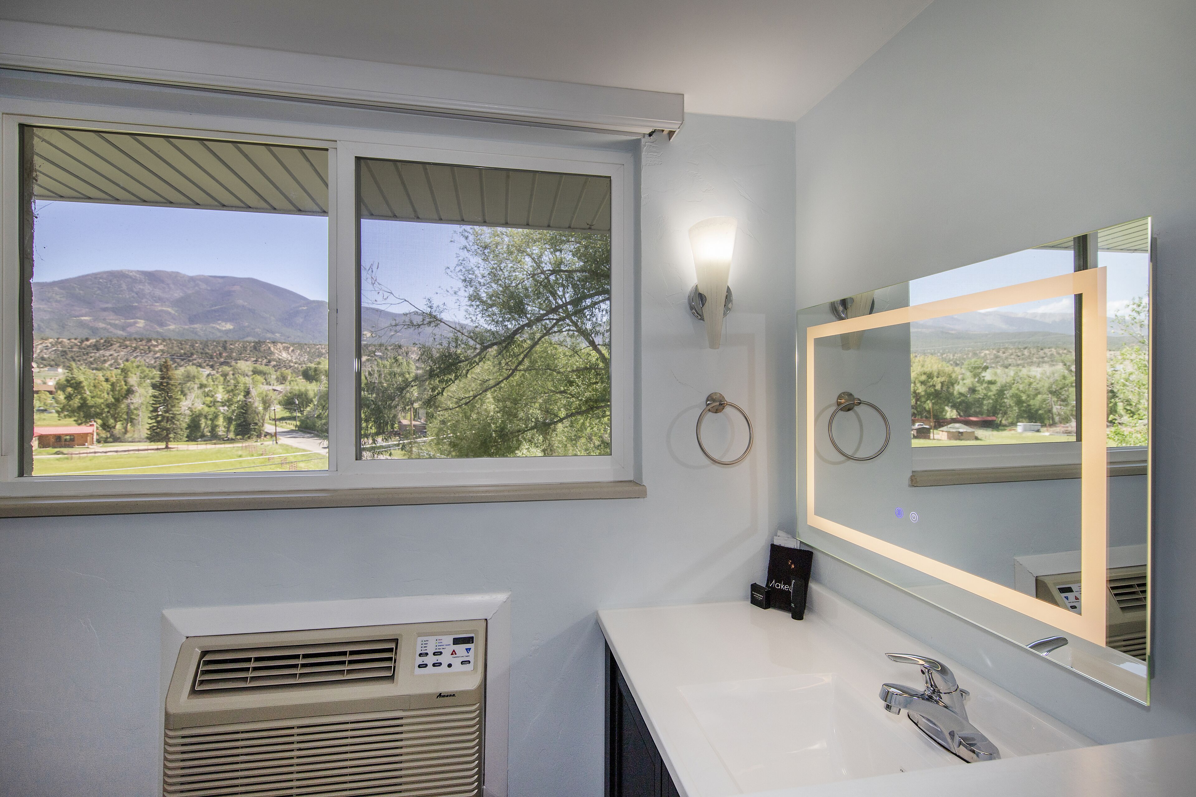 A bathroom with a large window, a wall-mounted heater, a sink, and a mirror with a light.