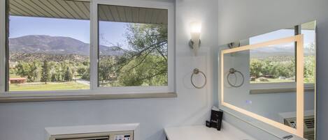 A bathroom with a large window, a wall-mounted heater, a sink, and a mirror with a light.