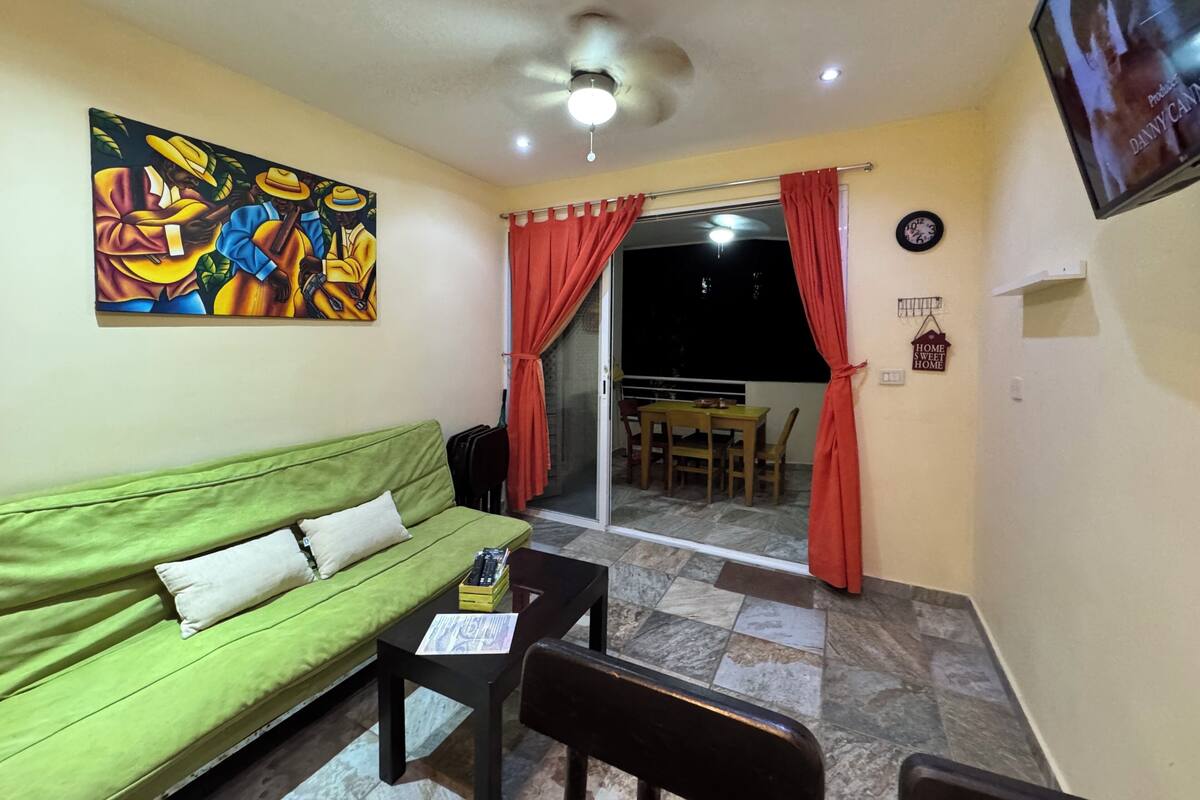 A living room with a green sofa, a small table, a ceiling fan, and a painting on the wall.