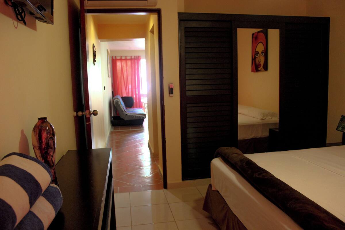 A hotel room with a bed, a desk, a chair, and a door leading to another room.