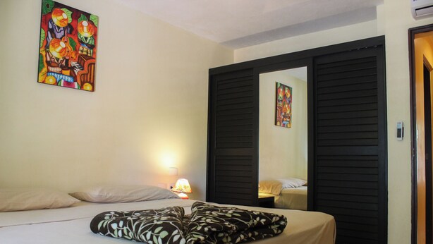 A hotel room with a bed, a nightstand with a lamp, a painting on the wall, and a door with a mirror. - Viva Maria Apartments (Playa del Carmen)