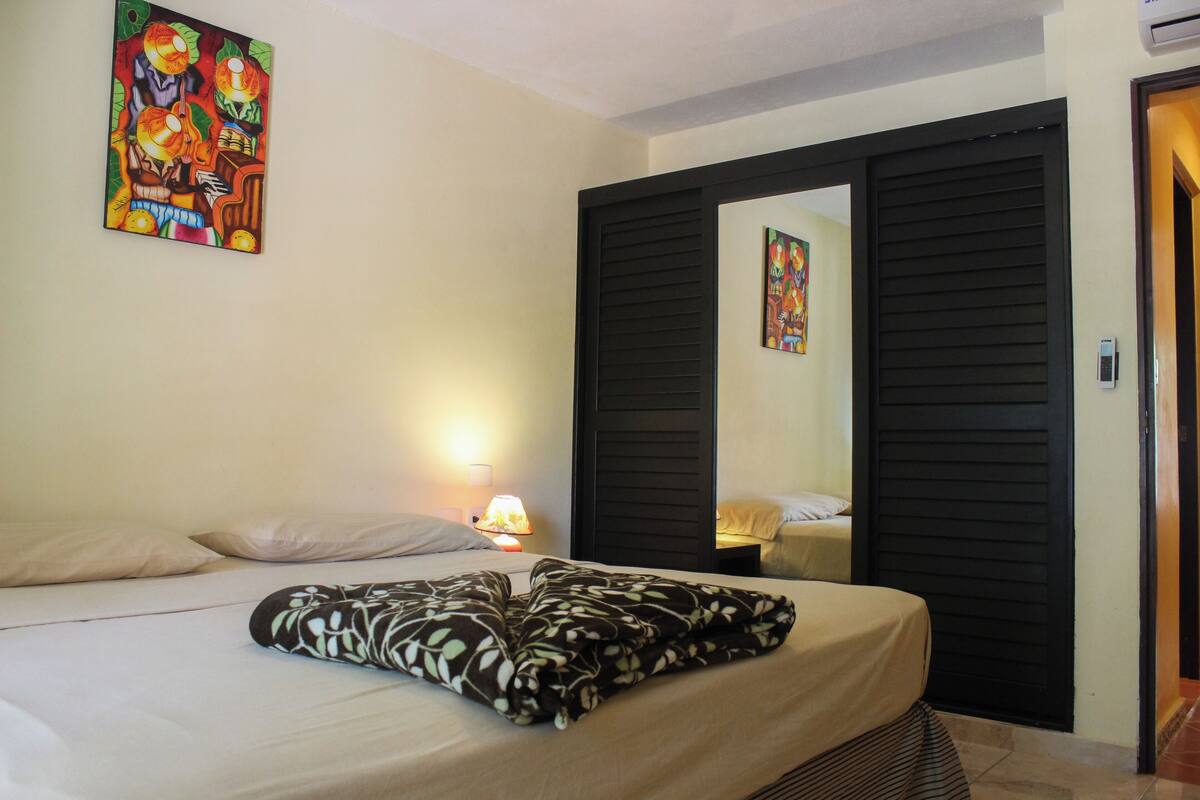 A hotel room with a bed, a nightstand with a lamp, a painting on the wall, and a door with a mirror.