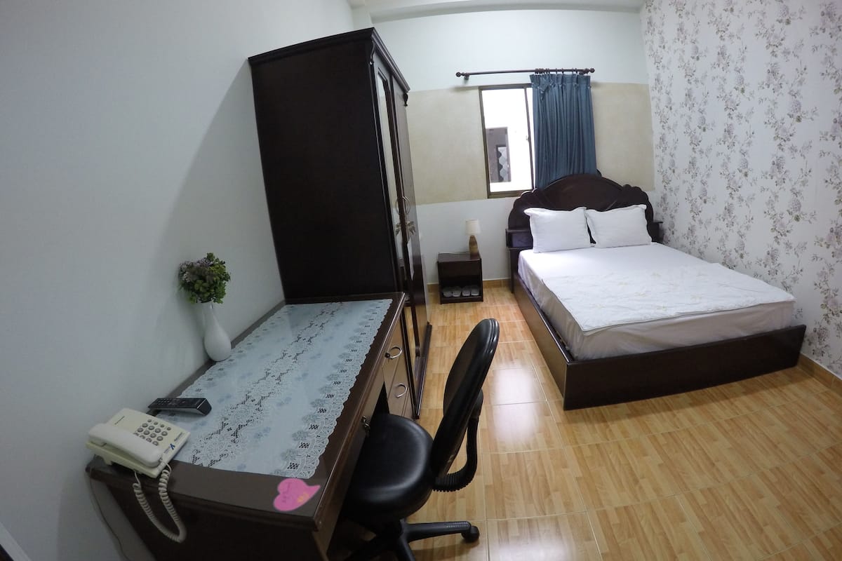 Ein Schlafzimmer mit einem Bett, einem Schreibtisch mit Telefon, einem Stuhl, einem Kleiderschrank, einem Nachttisch mit Lampe und einem Fenster mit Vorhängen.