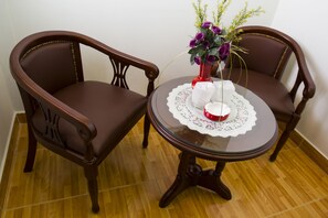 Two brown upholstered chairs with a glass table between them, a white lace tablecloth, and a vase with purple flowers.