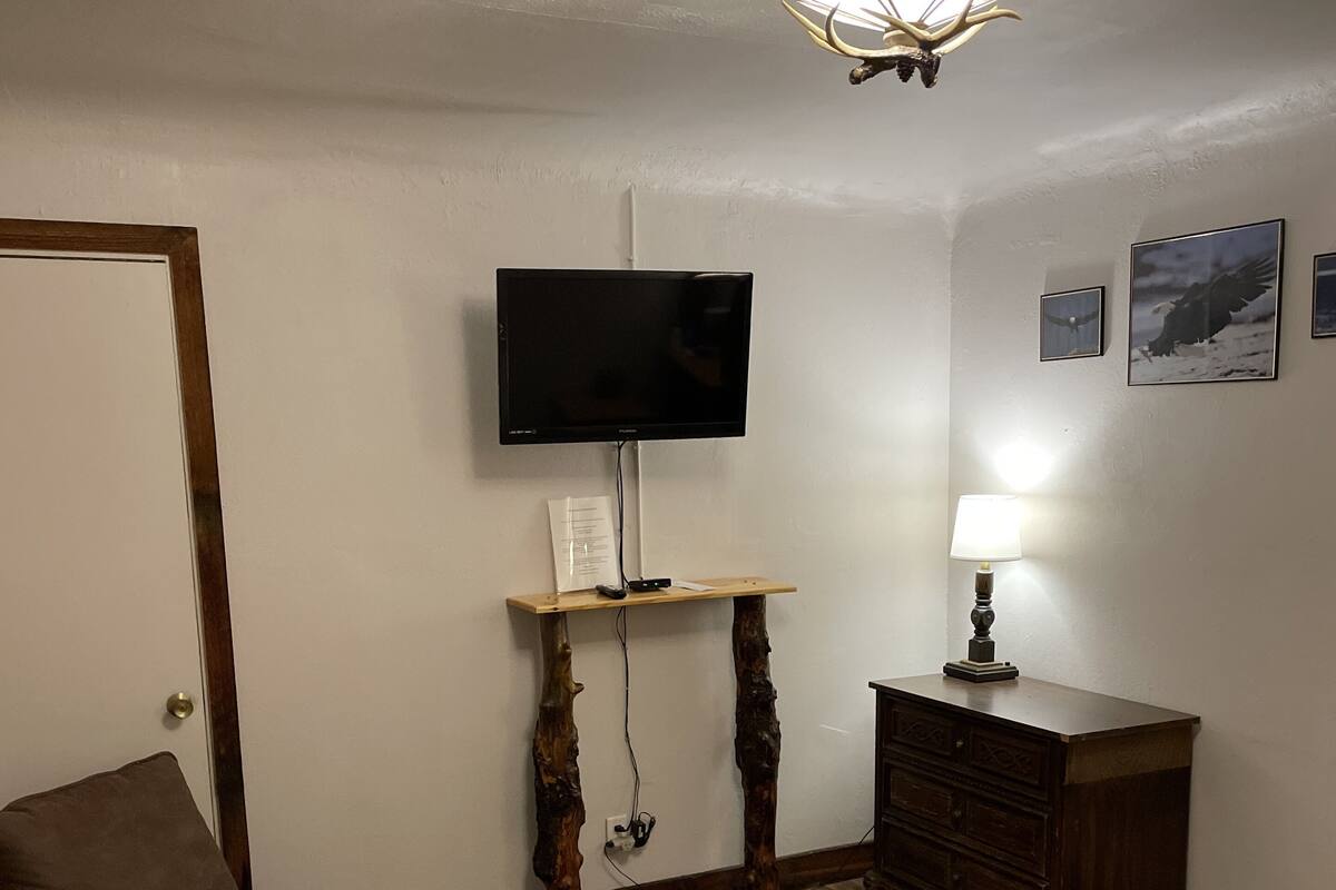 A bedroom with a bed, a chair, a TV on a wooden stand, a dresser, and a wall-mounted lamp.