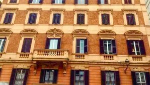 En bygning med flere etager og en symmetrisk facade, kendetegnet ved jævnt placerede vinduer med lukninger, en central balkon med gelændere og en ensartet arkitektonisk stil.