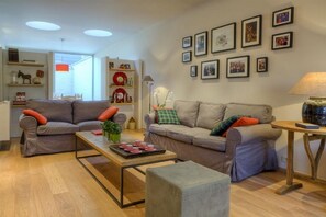 A living room with two sofas, a coffee table, and a lamp. There are framed pictures on the wall and shelves with decorative items.