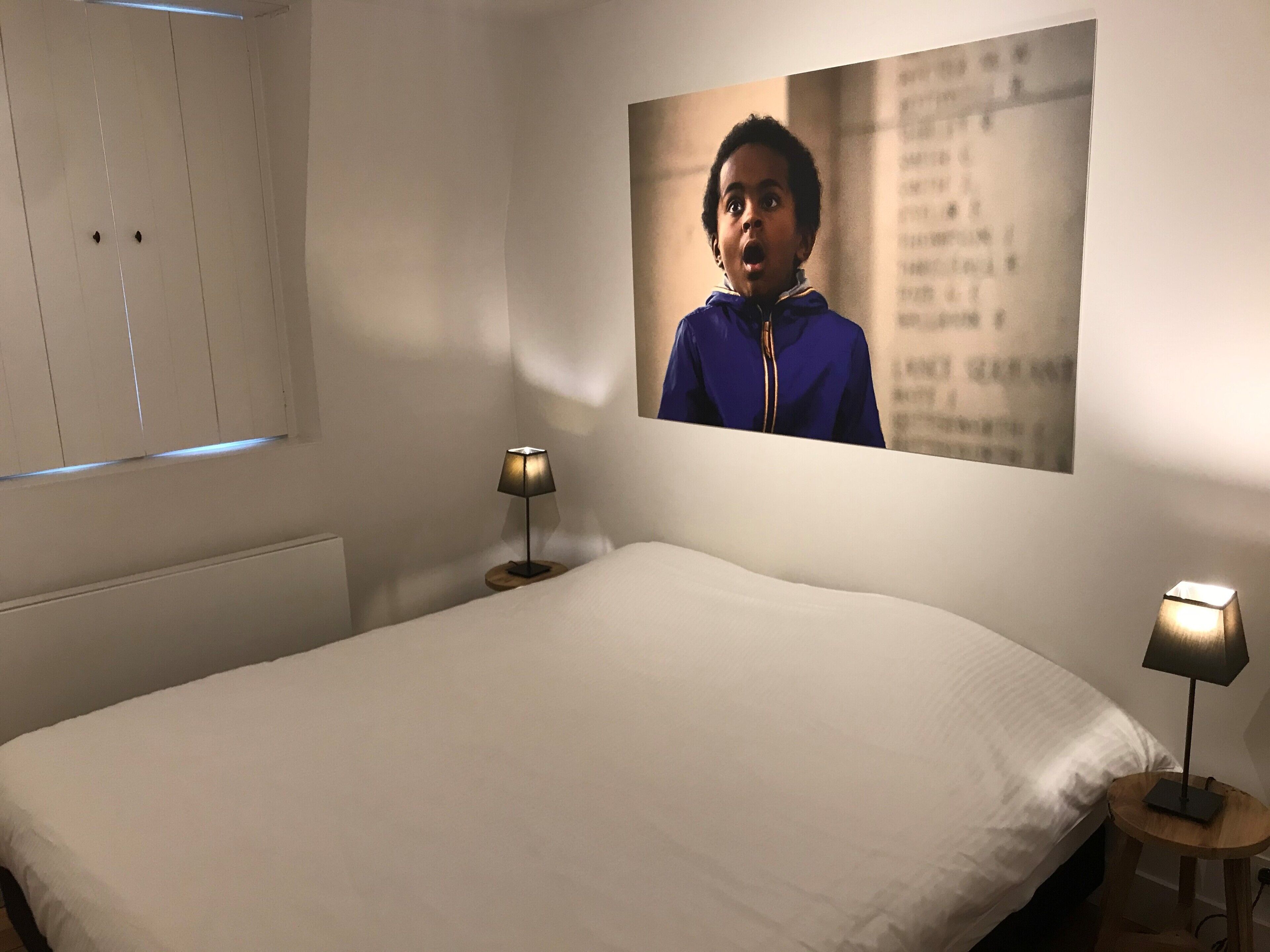 Een slaapkamer met een bed, twee bedlampjes, een raam met jaloezieën en een aan de muur hangend schilderij.