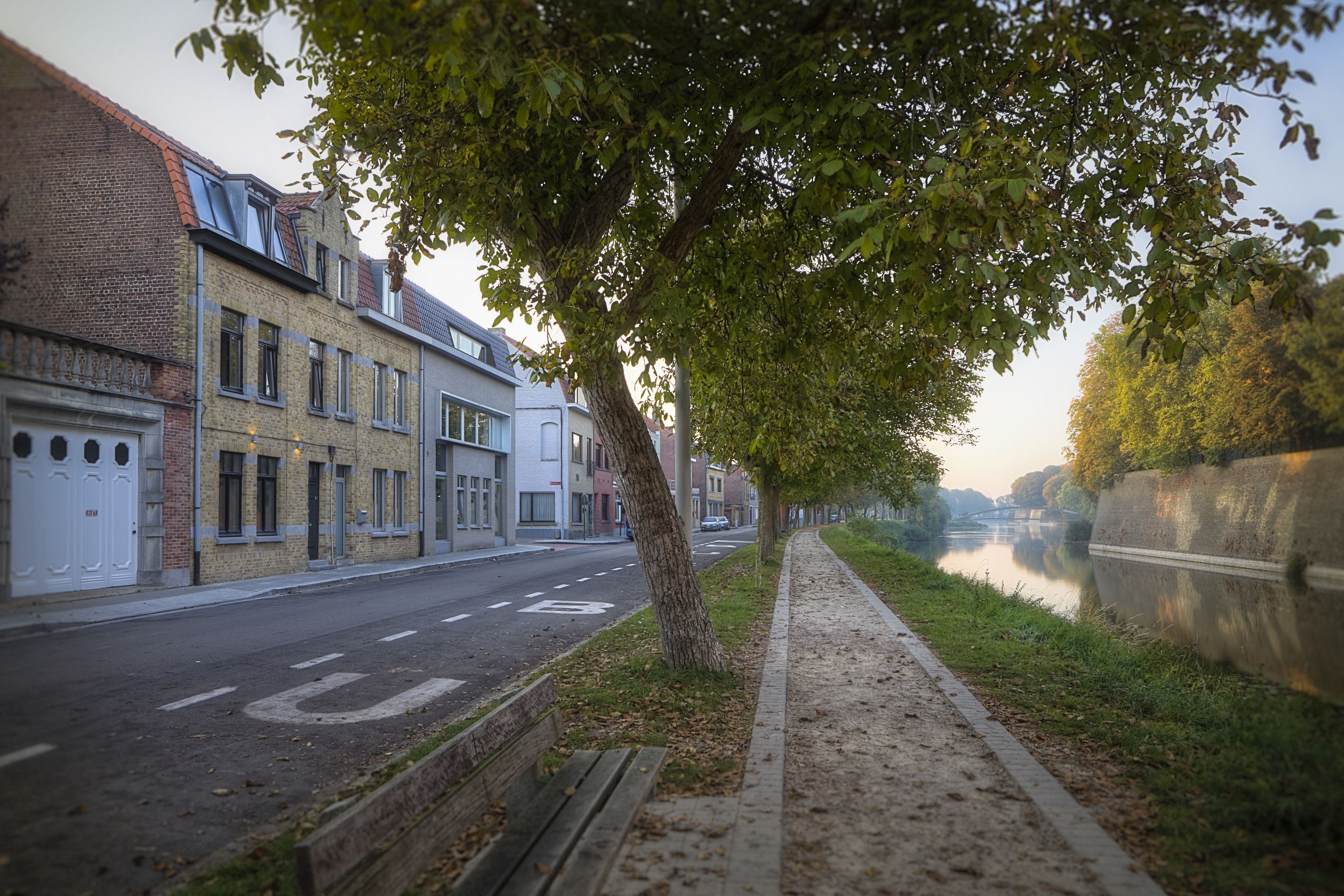 A tree-lined street with a canal, buildings, and a bench.
