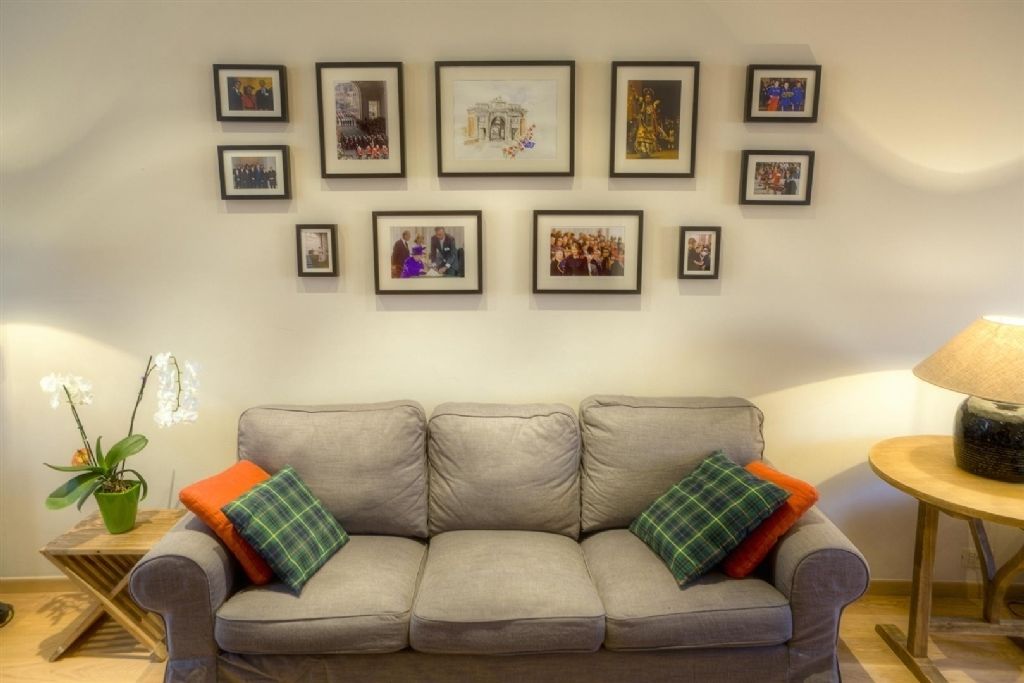A living room with a grey sofa, framed pictures on the wall, a small table with a lamp, and a potted plant.