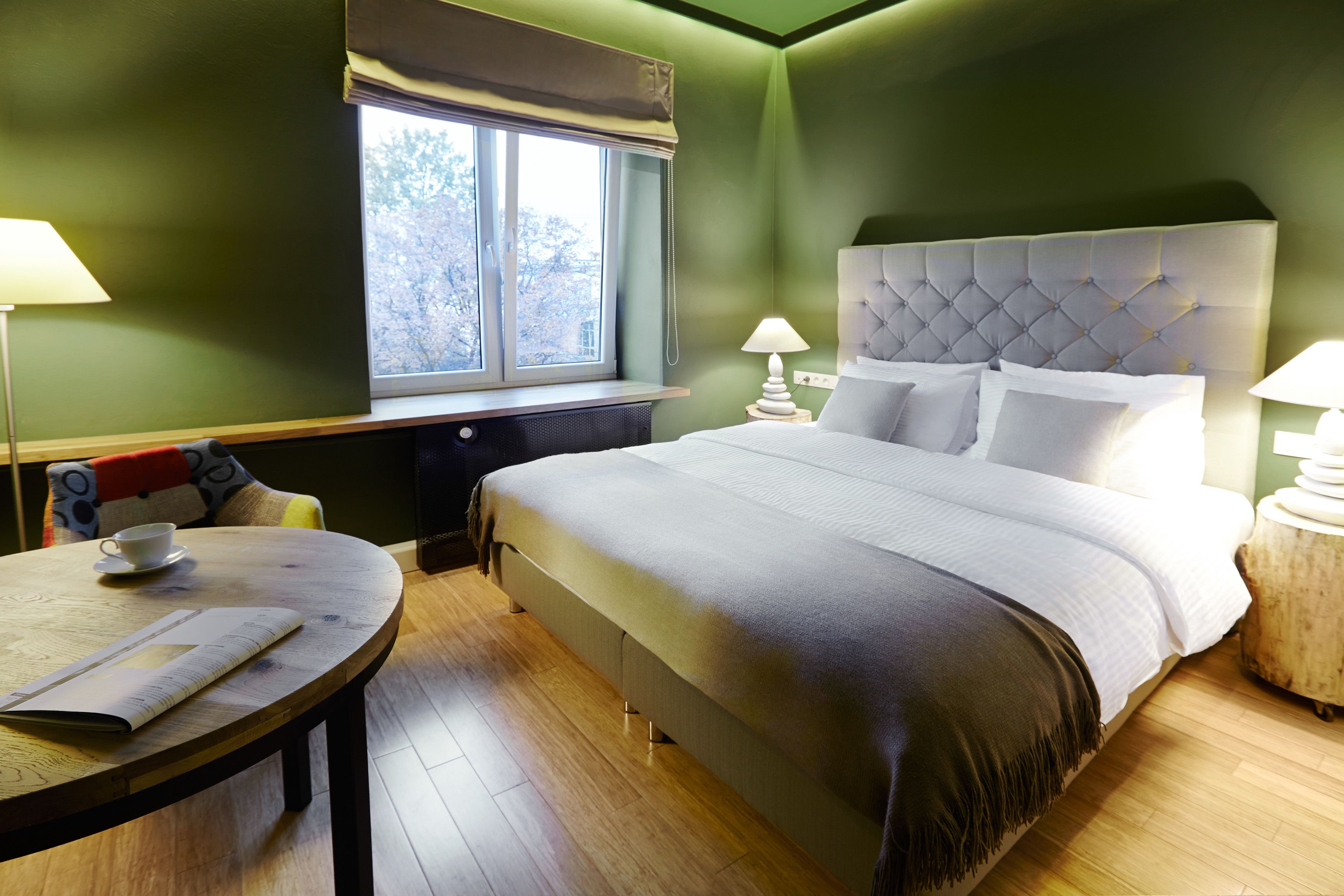 A hotel room with a large bed, two bedside tables with lamps, a round wooden table with a cup and menu, and a window with a view of trees.