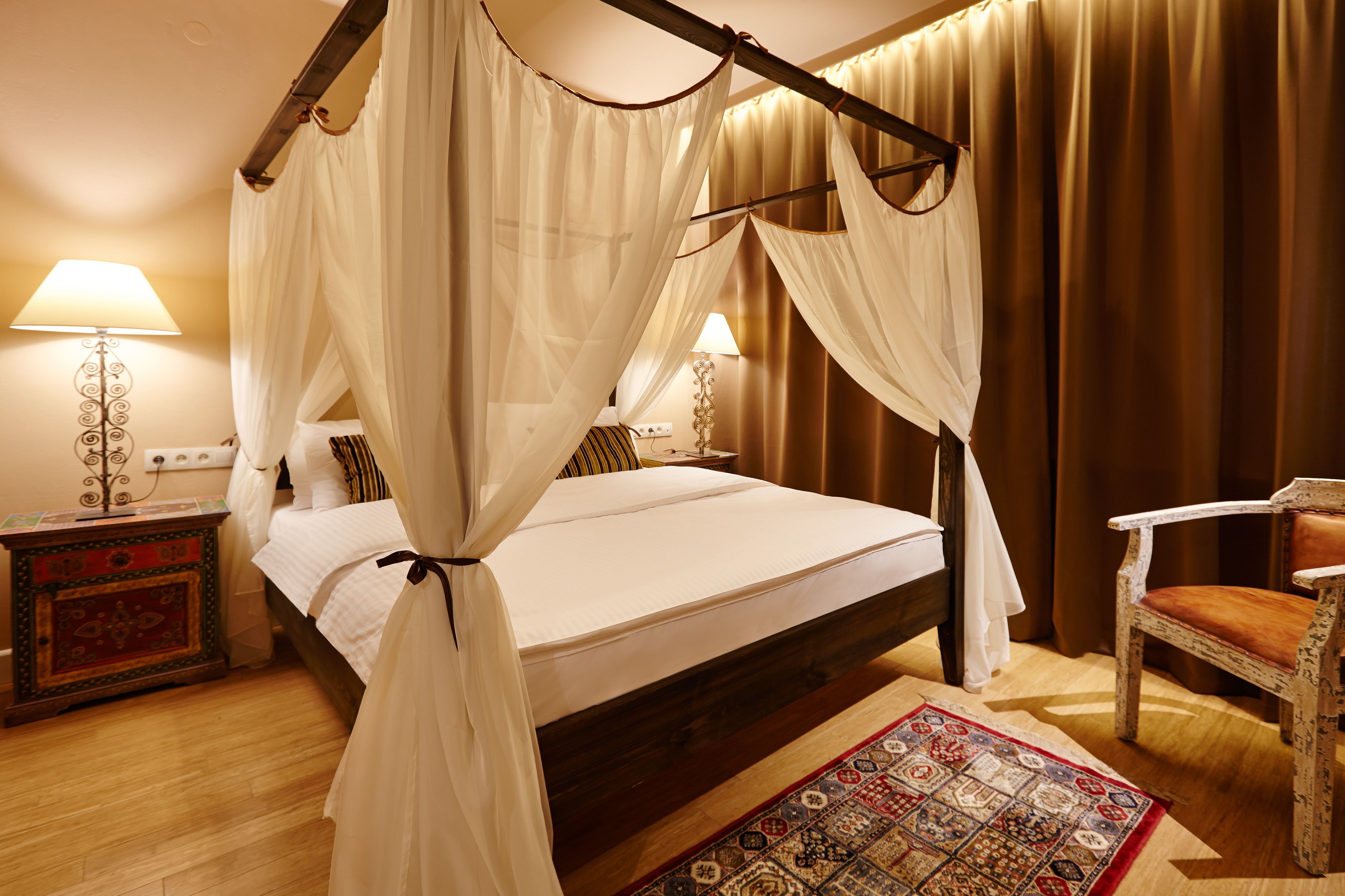 Un lit à baldaquin à quatre poteaux avec une structure en bois, une table de chevet avec une lampe, un tapis à motifs et une chaise en bois.