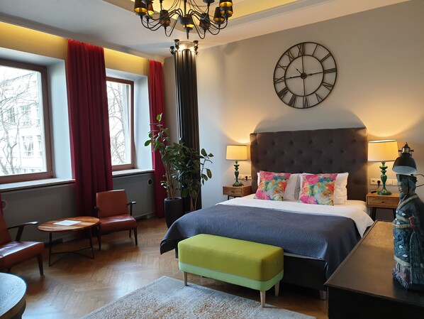 Habitación de hotel con una cama grande, un candelabro, un reloj, una planta y vistas al exterior.