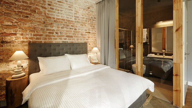 A bedroom with a brick wall, a large bed, two bedside lamps, and a sliding glass door leading to a bathroom. - SleepWell Apartments Ordynacka (Warsaw)